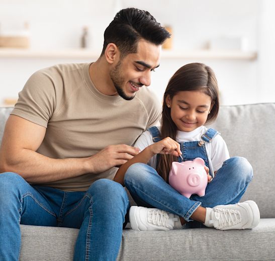A father sitting beside his daughter who is putting a coin into a piggy bank.