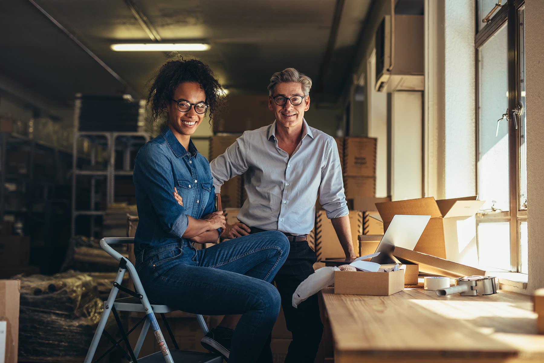 Two people sitting in a storage area with boxes, having a conversation in natural light.