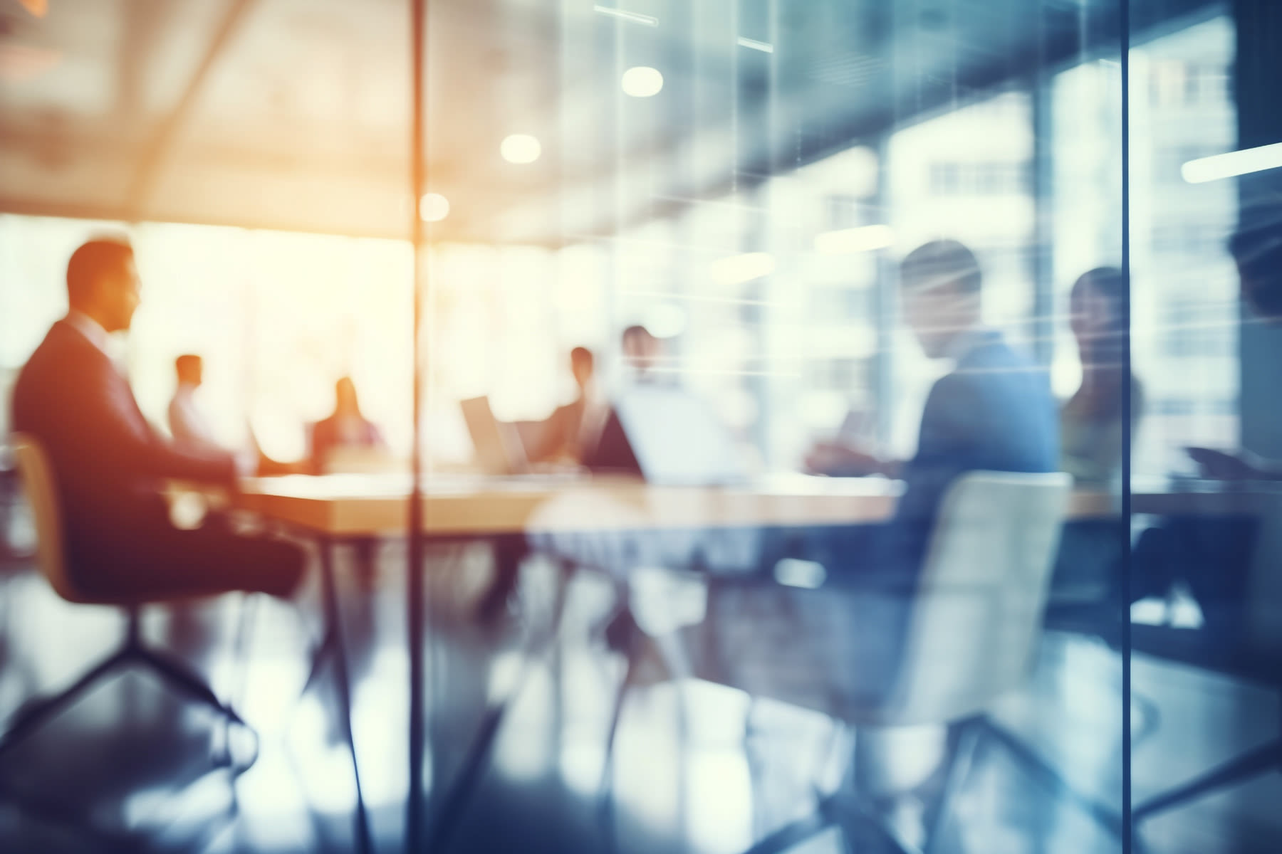 A blurred image of professionals in a meeting room with glass walls, illuminated by natural light.