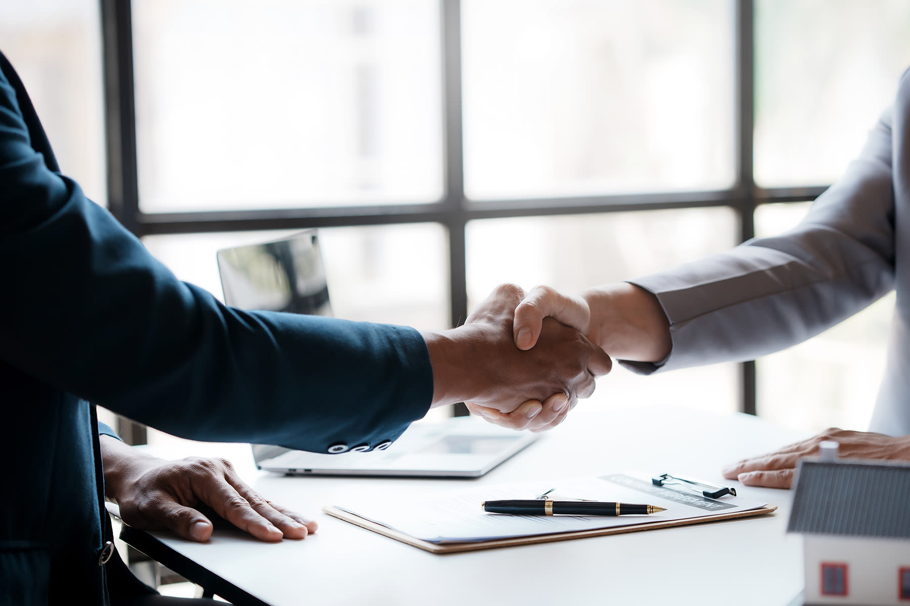 Two individuals shaking hands across a table with documents, a pen, and a model house, indicating a property deal or agreement.