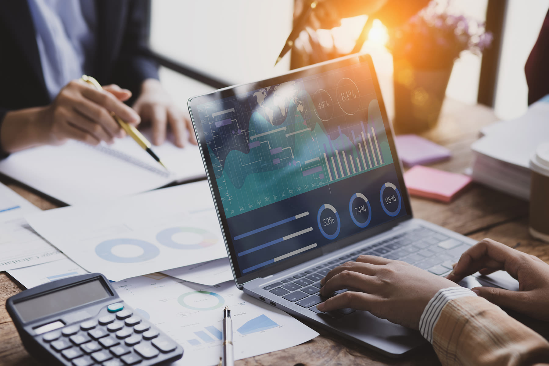 Two people working at a desk and one is typing on a computer and the other is pointing at some papers on the desk