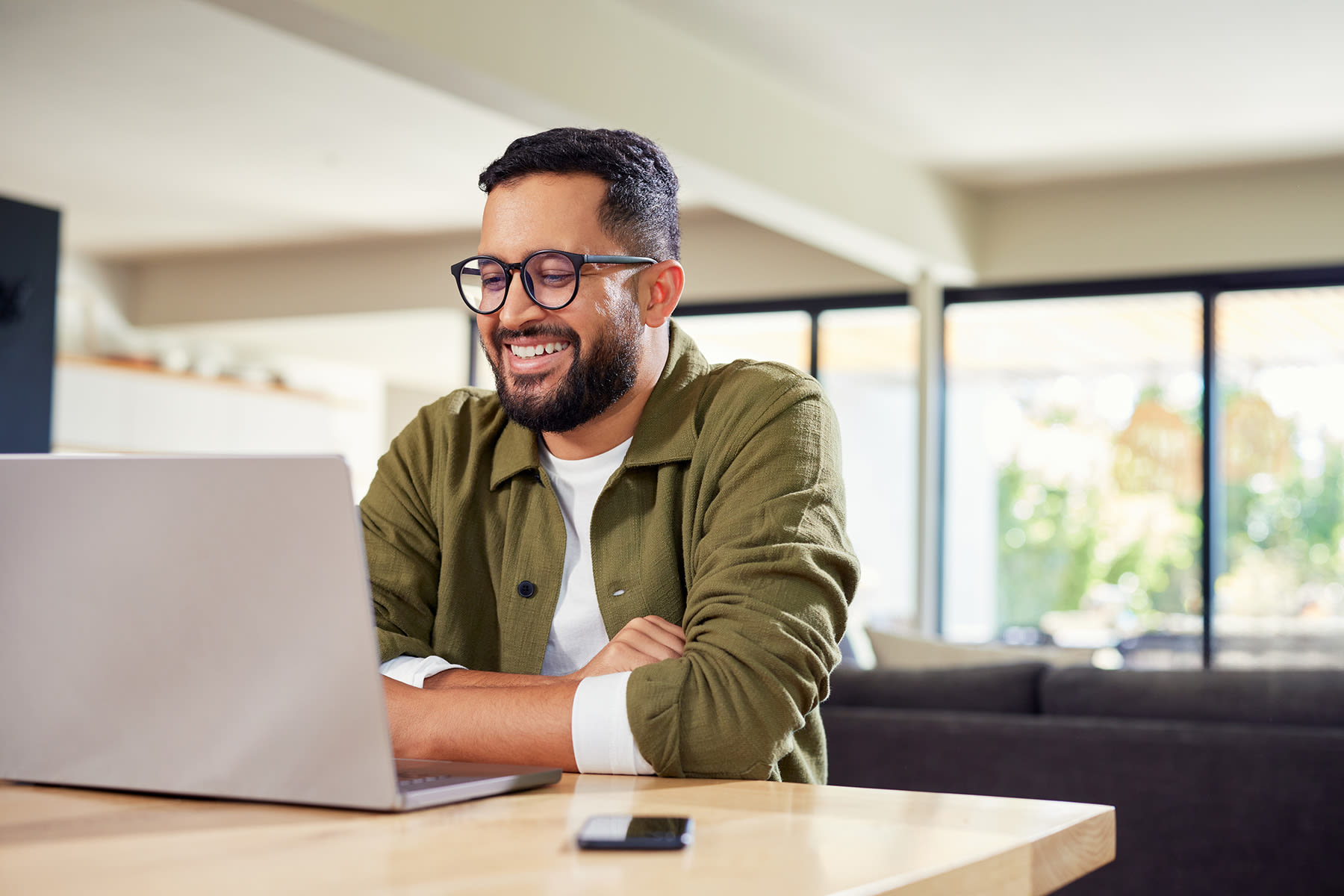 Person with short hair and glasses using a laptop at a wooden table in a bright, modern living space.