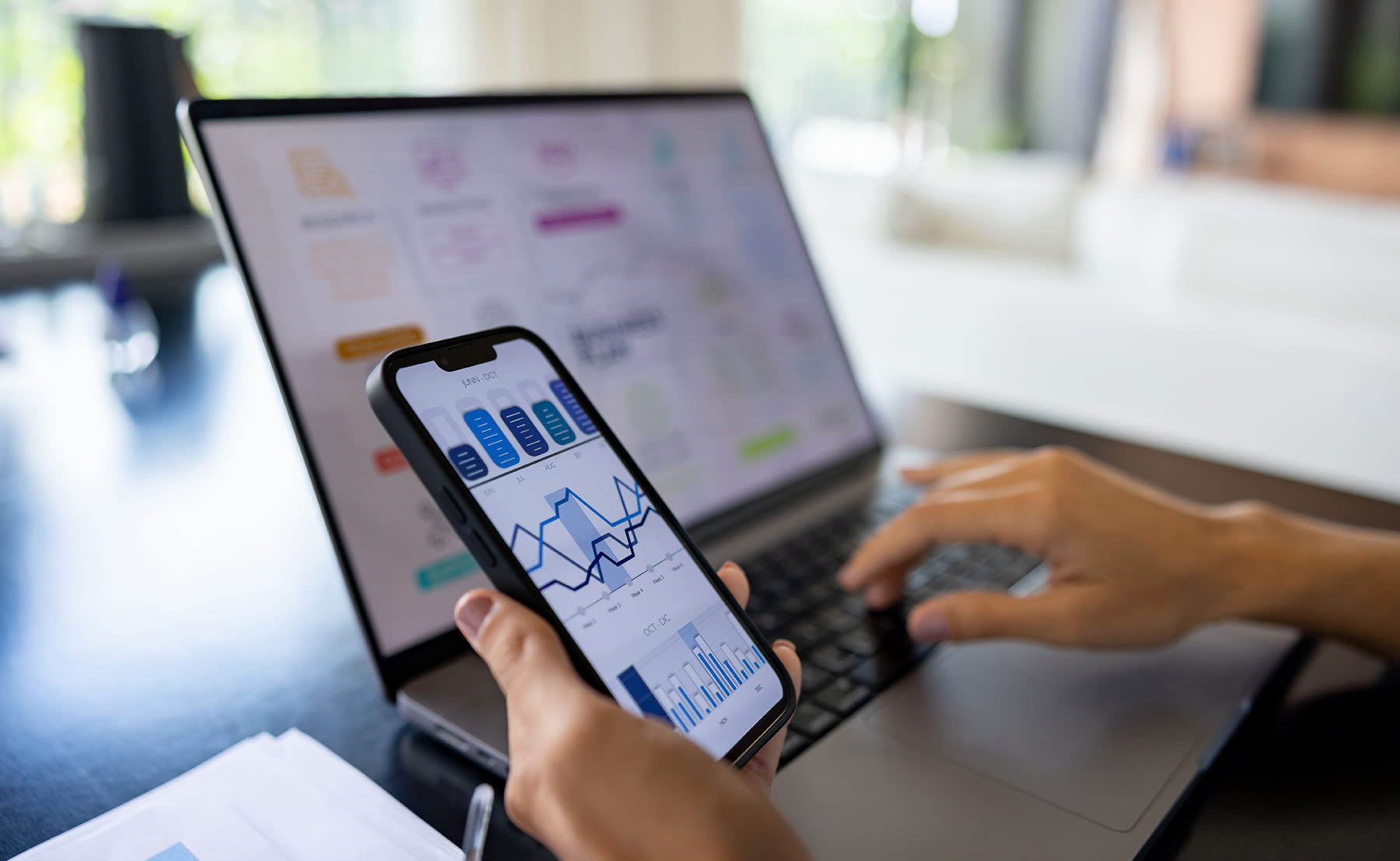 Person using a laptop and holding a smartphone displaying charts and graphs