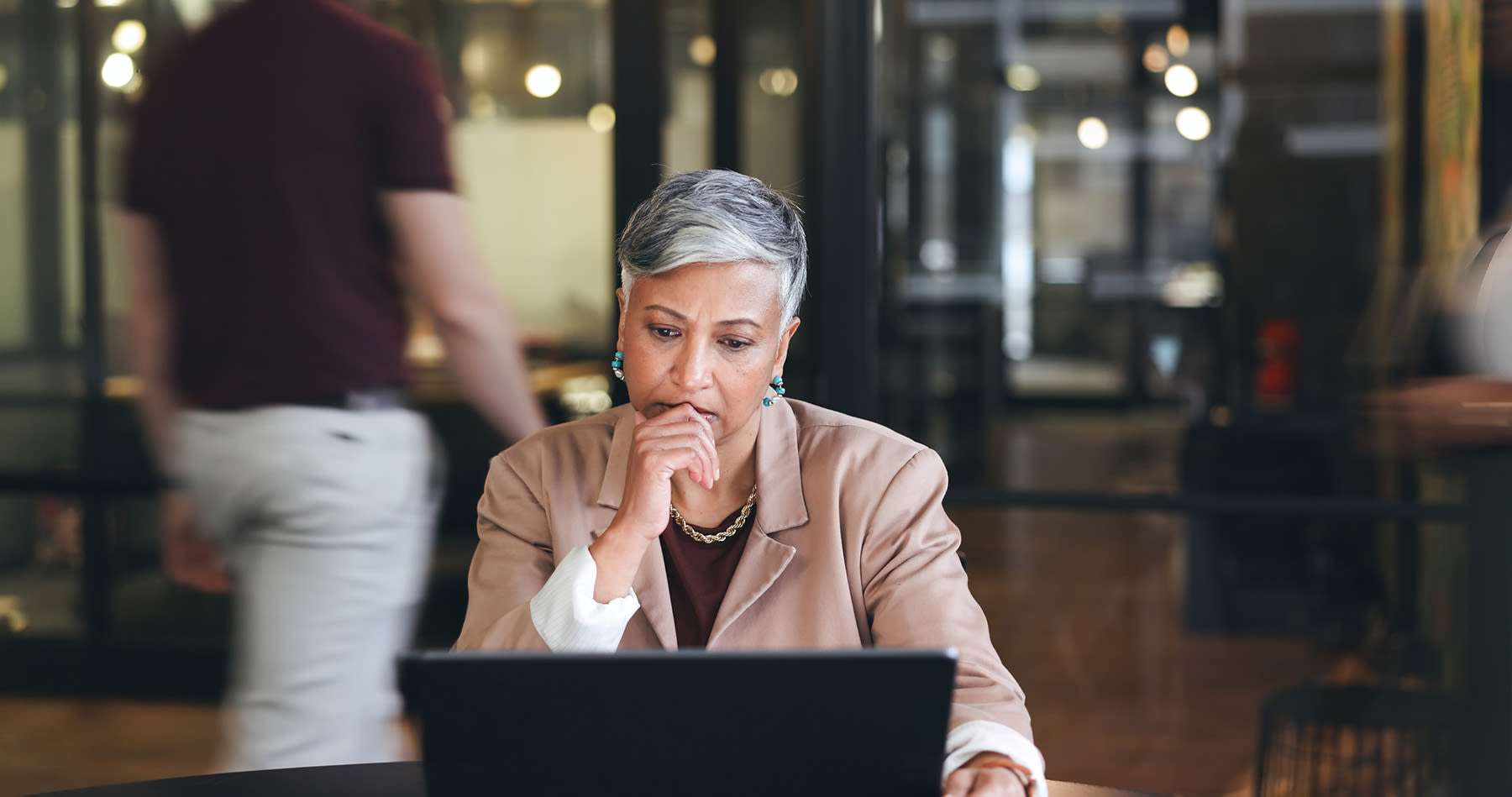 A person with short gray hair, wearing a beige blazer and white shirt, sits at a desk working on a laptop in an office with glass walls and blurred figures in the background.