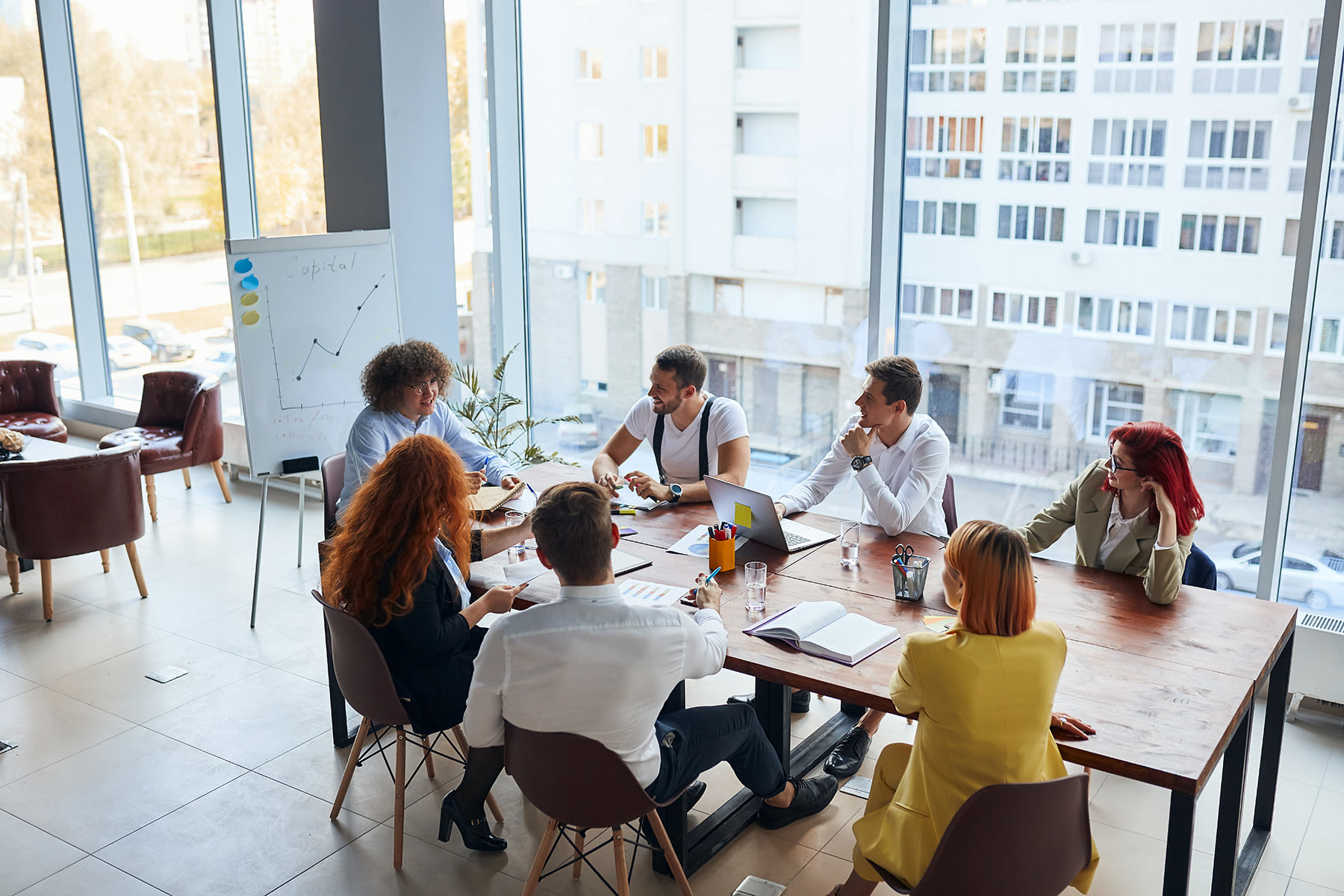 A group of professionals is engaged in a meeting around a wooden table, discussing ideas with notebooks and laptops open, in a bright room with large windows overlooking the city.