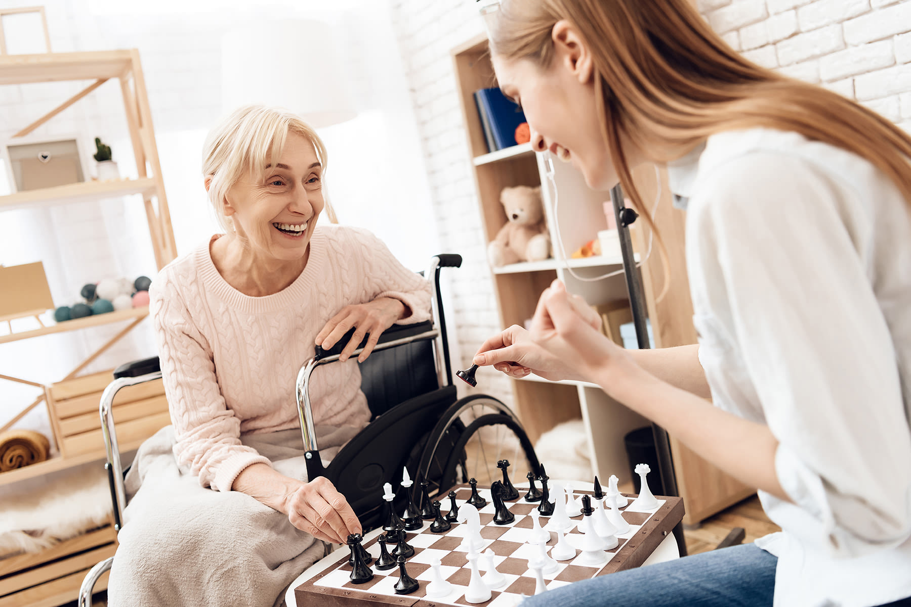 An elderly woman seated in a wheelchair playing chess with a younger woman.