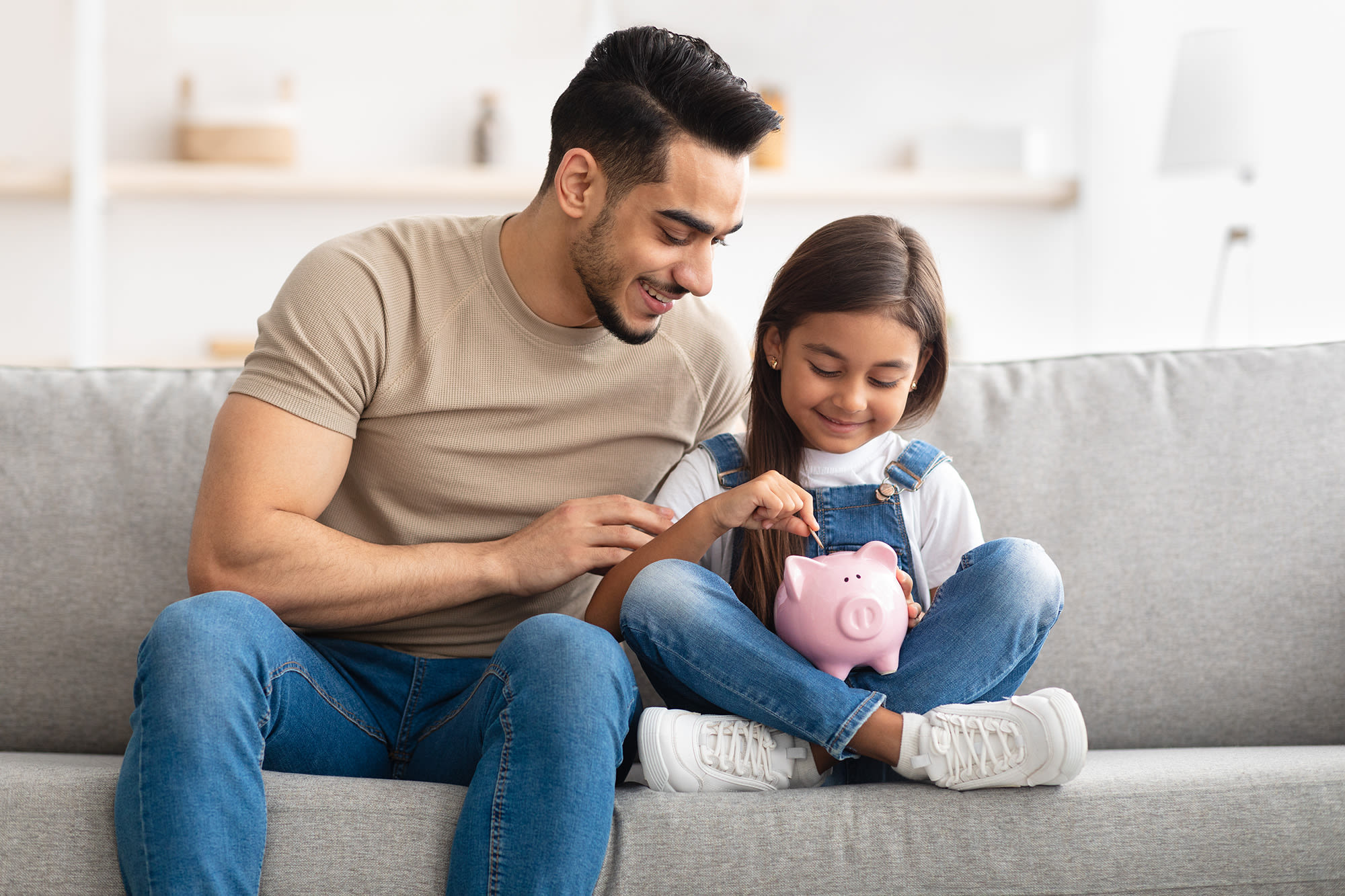 A father sitting beside his daughter who is putting a coin into a piggy bank.