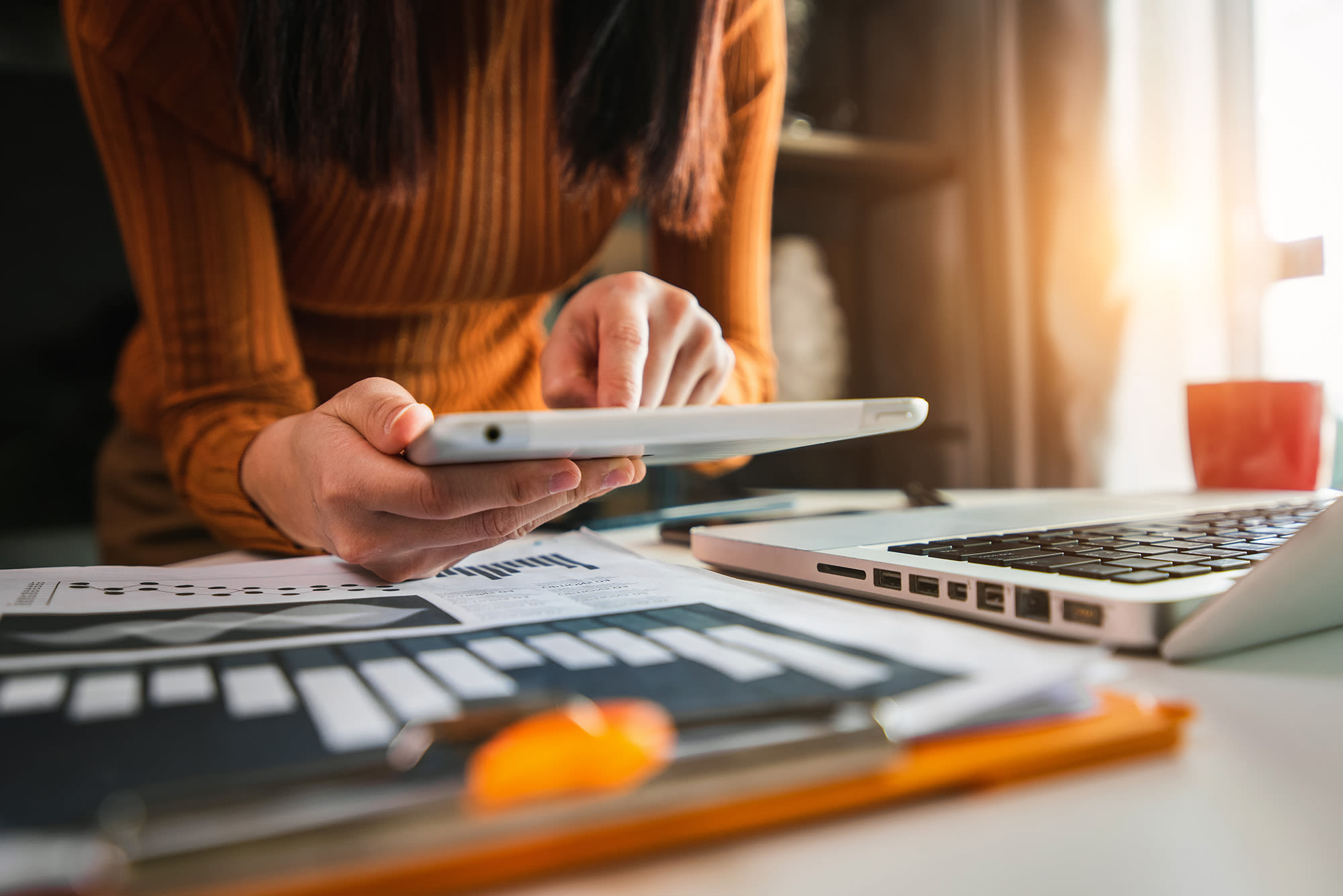 a person in an orange sweater working at a desk holding a tablet