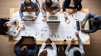 A top-down view of a group of professionals collaborating around a table, working on laptops and analyzing various charts and graphs printed on papers.