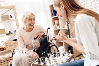 An elderly woman seated in a wheelchair playing chess with a younger woman.