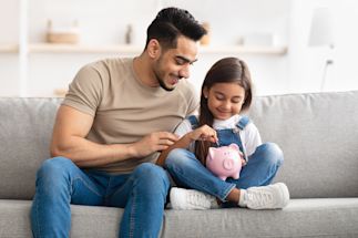 A father sitting beside his daughter who is putting a coin into a piggy bank.