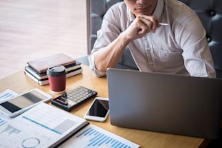 Man seated at a desk with a laptop computer, phone, calculator and other business equipment.