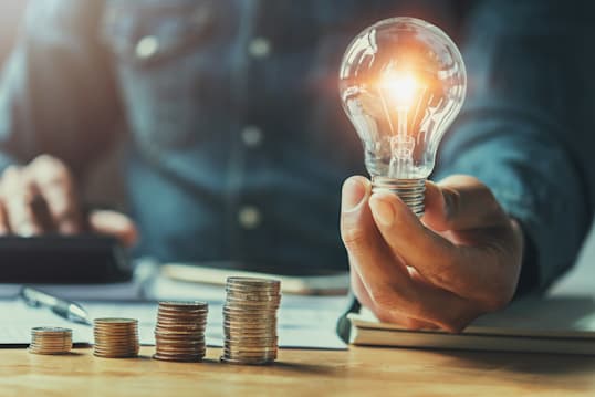 person holding a lightbulb with a stack of coins on a desk