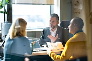 3 individuals sitting at a table discussing and looking at a laptop