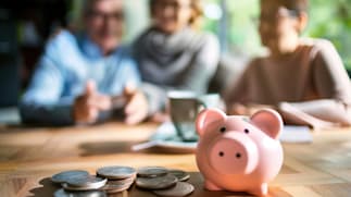 A pink piggy bank on a wooden table with scattered coins, with three blurred individuals in the background.