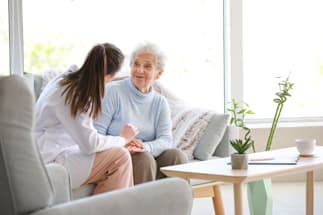 A healthcare professional comforts an elderly person on a couch, with their hand on the elderly person’s back. Faces are obscured for privacy.