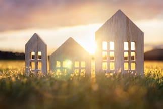 Three wooden house-shaped cutouts are placed on a surface with the sun setting in the background. The sunlight shines through the windows of the middle house cutout, creating a warm glow.